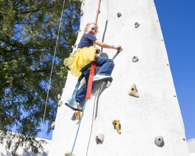 An HRA student smiling while ascending a rock climbing wall at the 2025 HRA Fall Festival