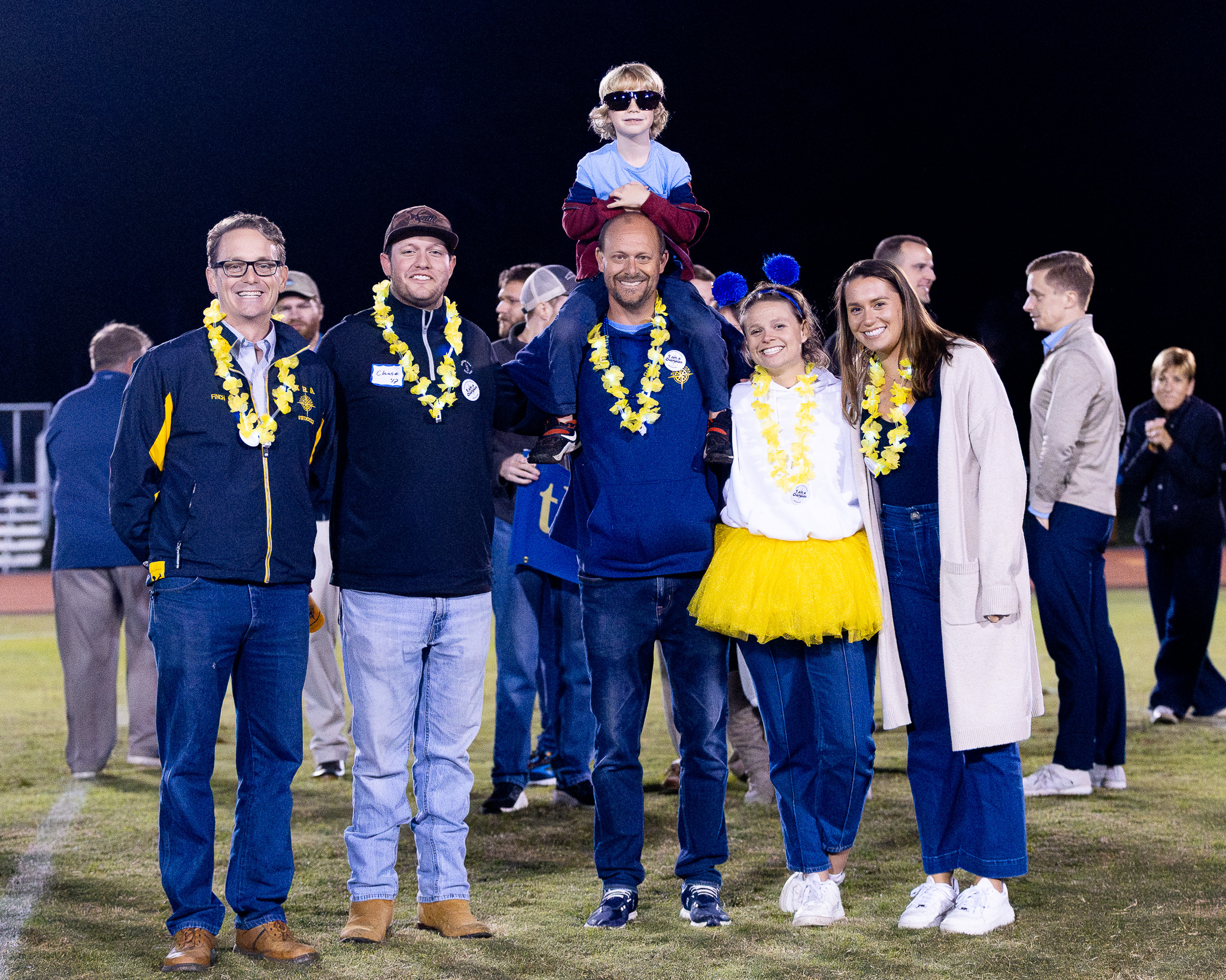 HRA varsity swimming coach Jeff Scott with alumni and former swimmers Mitchell Finch '08, Chase Suttle '17, Caroline Thatcher '16, and Peyton Baldwin '15, as well as his son Patrick, on the football field at Homecoming 2025