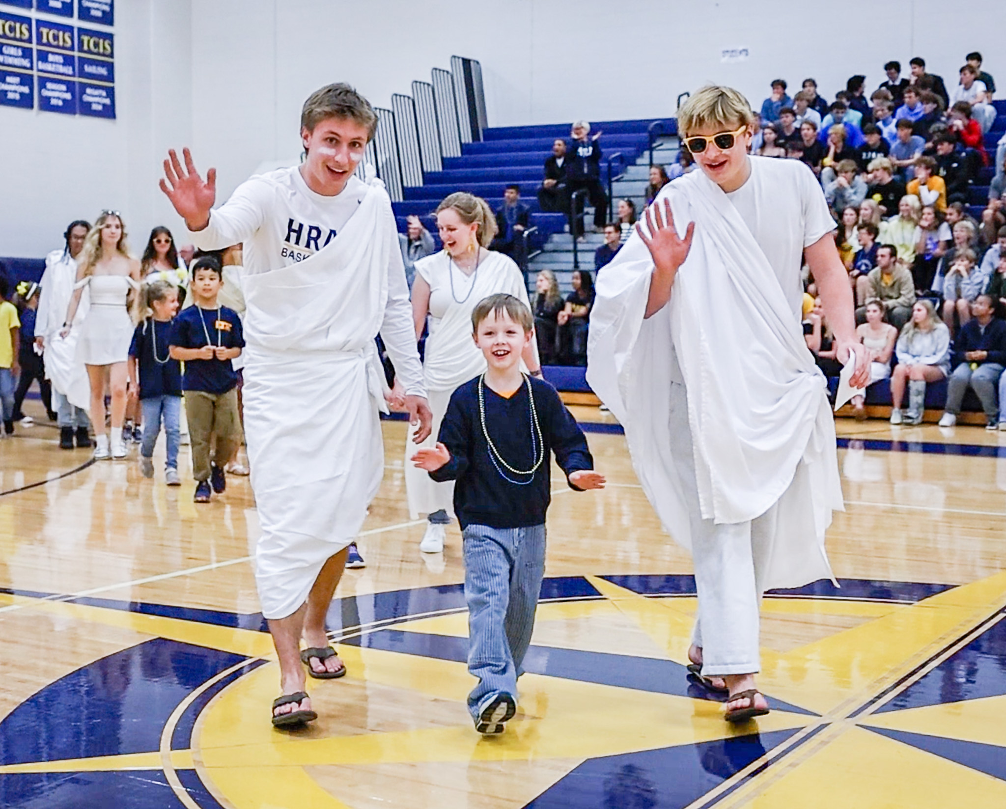 Two HRA seniors in togas in the gymnasium with their first-grade buddy