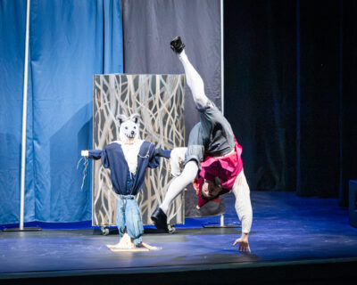 An actor performing a front flip in front of a Big Bad Wolf doll during Virginia Opera's October 2026 production of "The Three Little Pigs" for HRA's Lower School