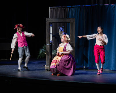 Two actors pointing and laughing at a seated actress with a book during Virginia Opera's October 2026 production of "The Three Little Pigs" for HRA's Lower School